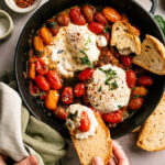 Overhead view of warm burrata with burst cherry tomatoes in a rustic skillet with toasted artisan bread being served by hand.