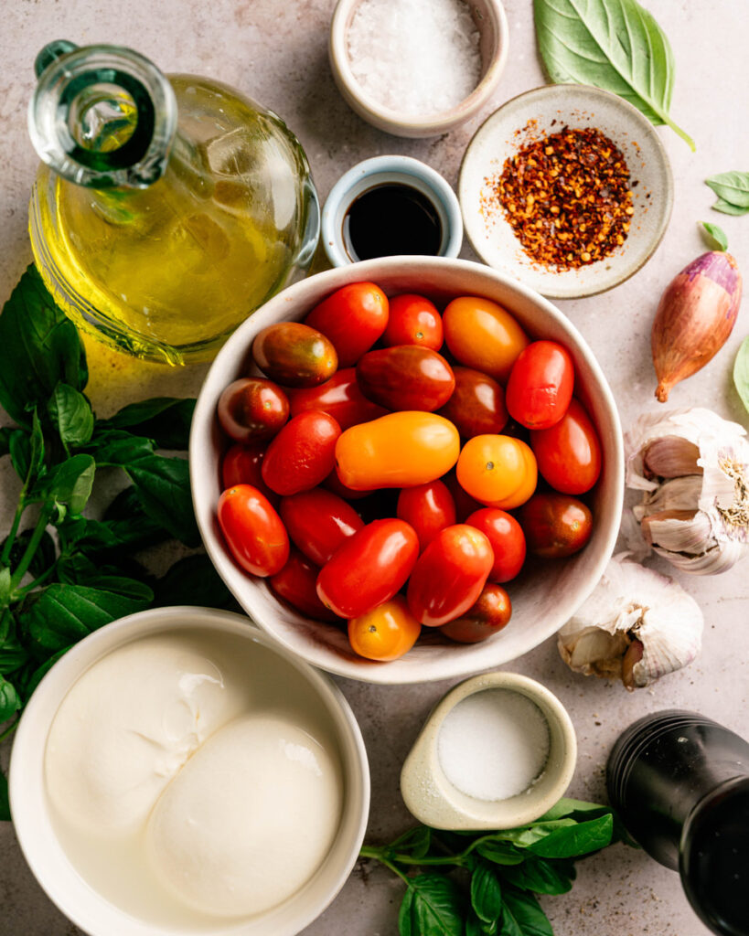 Fresh ingredients for warm burrata with burst cherry tomatoes, including burrata cheese, colorful cherry tomatoes, garlic, basil, olive oil, balsamic vinegar, sea salt, black pepper, and red pepper flakes arranged on a rustic surface.