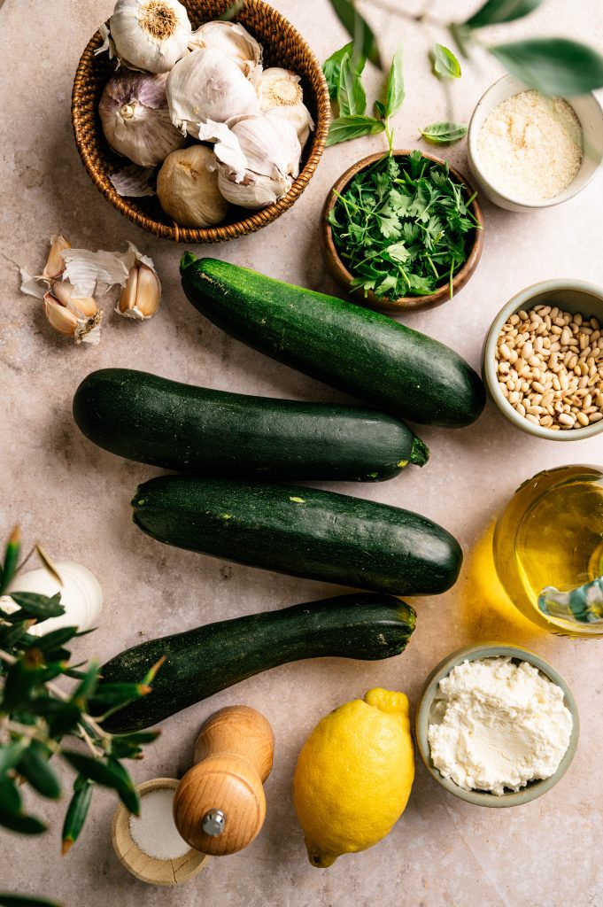 Ingredients for lemon ricotta zoodles arranged with zucchini, ricotta, lemon, herbs, garlic, Parmesan, pine nuts, and olive oil.