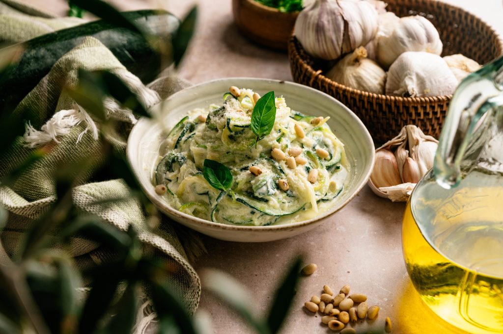 Creamy lemon ricotta zoodles served with basil and pine nuts beside garlic and olive oil.