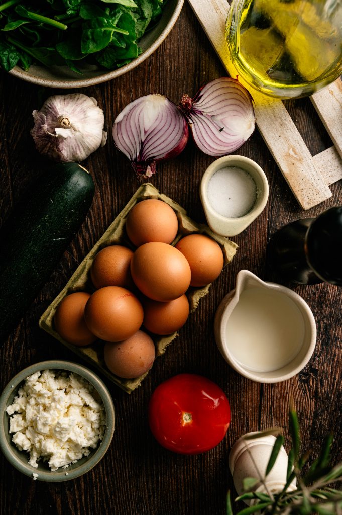 Ingredients for garden zucchini frittata arranged on a rustic table, including eggs, zucchini, tomato, spinach, onion, garlic, milk, cheese, and olive oil.