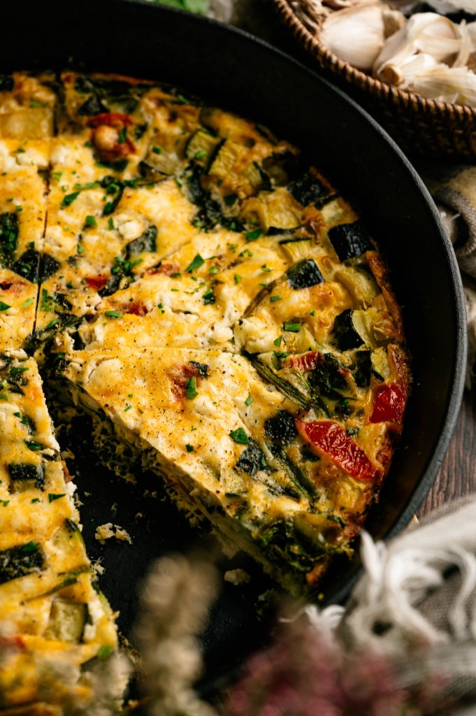 Garden zucchini frittata with a slice removed from a cast iron skillet, showing zucchini, tomatoes, spinach, and cheese.