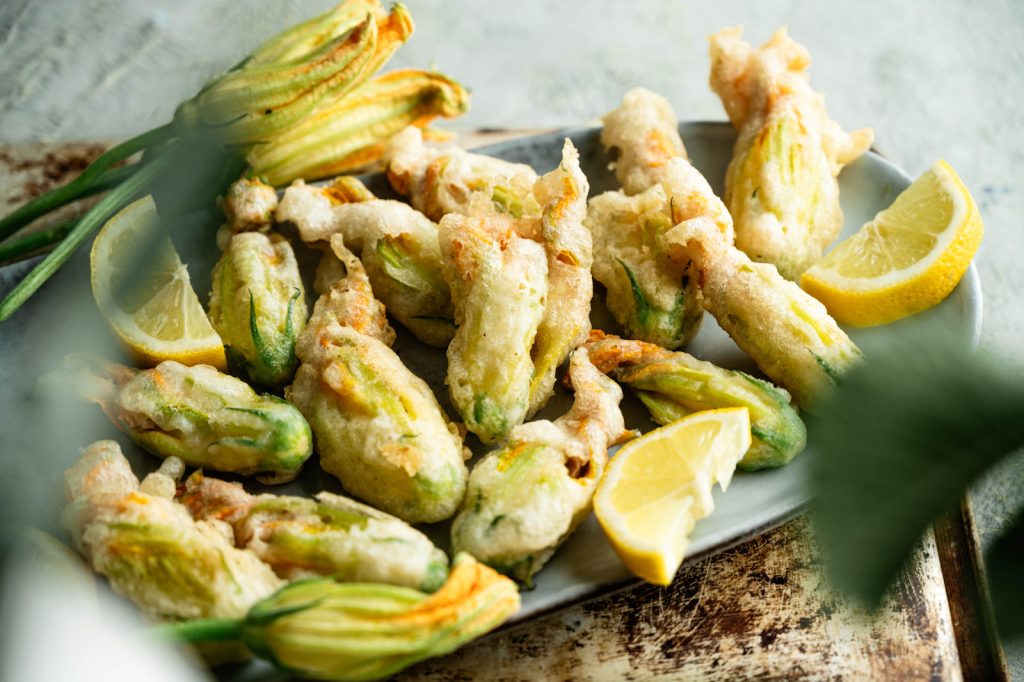 Italian fried zucchini blossoms piled on a gray platter with lemon and fresh blossoms
