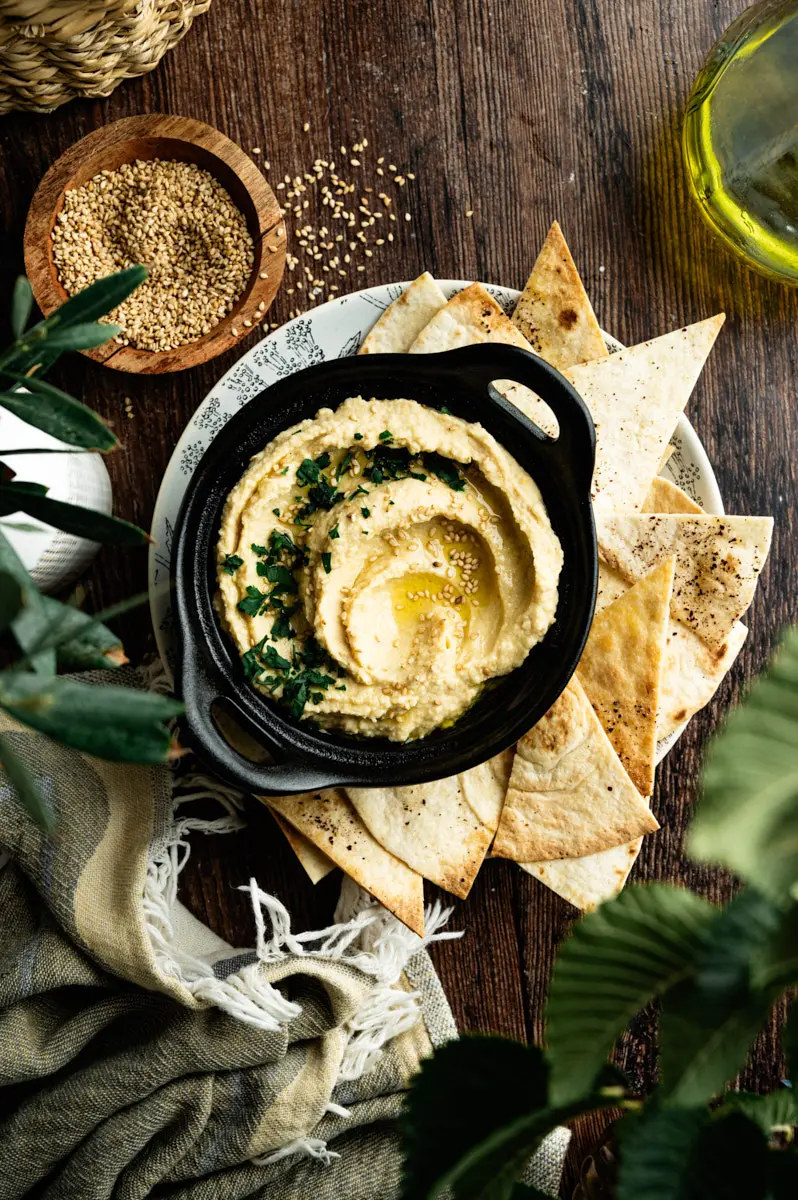 Overhead view of homemade hummus surrounded by pita bread and rustic linens