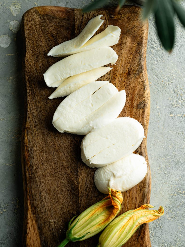Strips of fresh mozzarella on a wooden board beside two zucchini blossoms