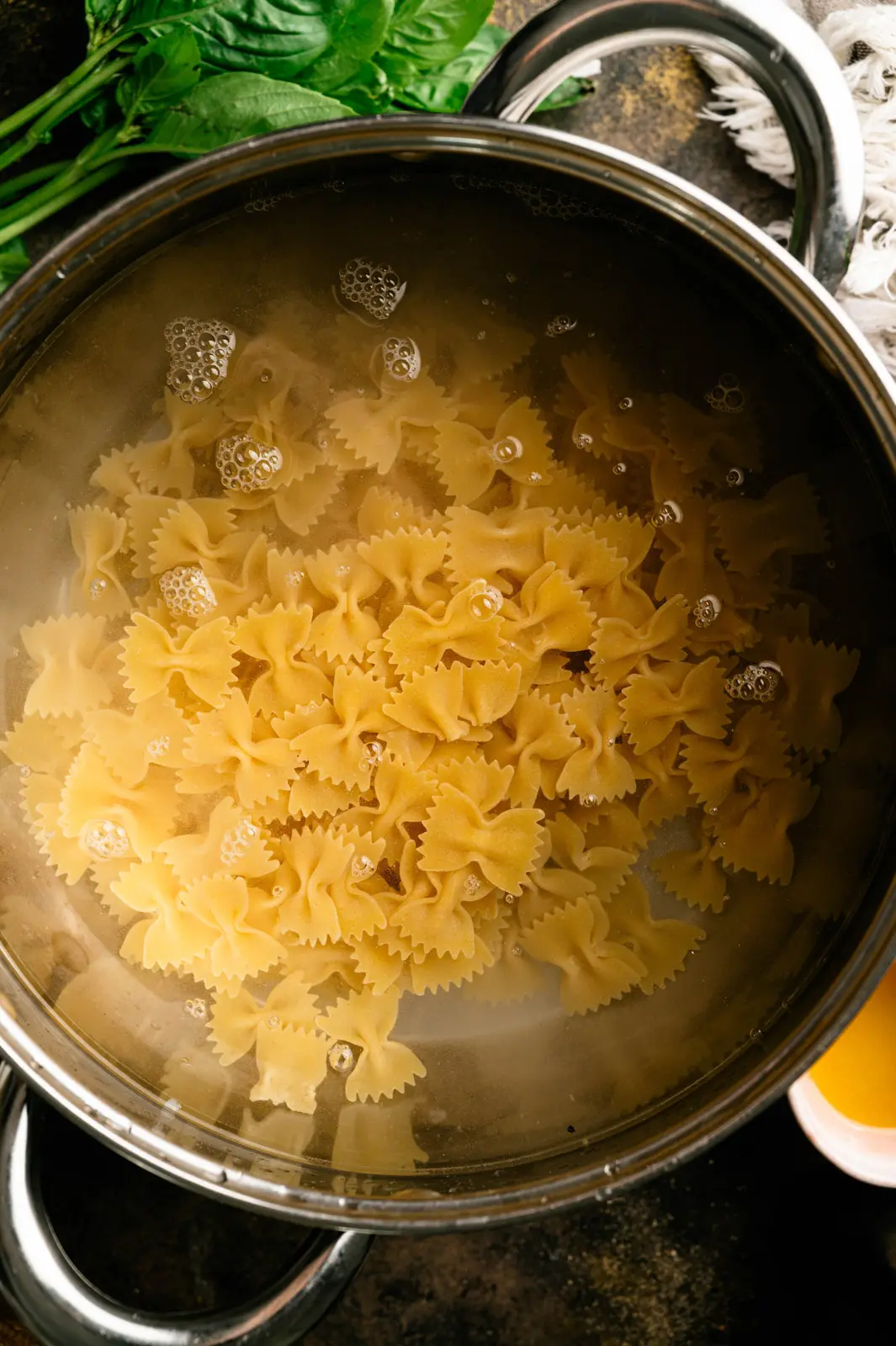 Farfalle pasta cooking in water in a stainless steel pot, with fresh basil beside it.