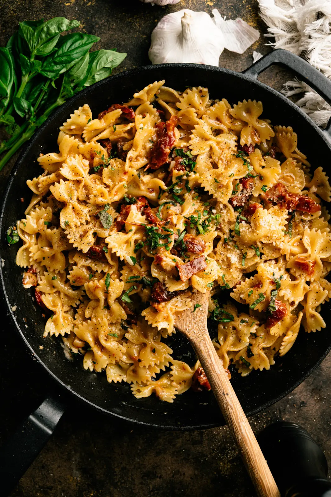 Sun-dried tomato and basil farfalle topped with grated cheese in a cast-iron skillet with a wooden spoon.