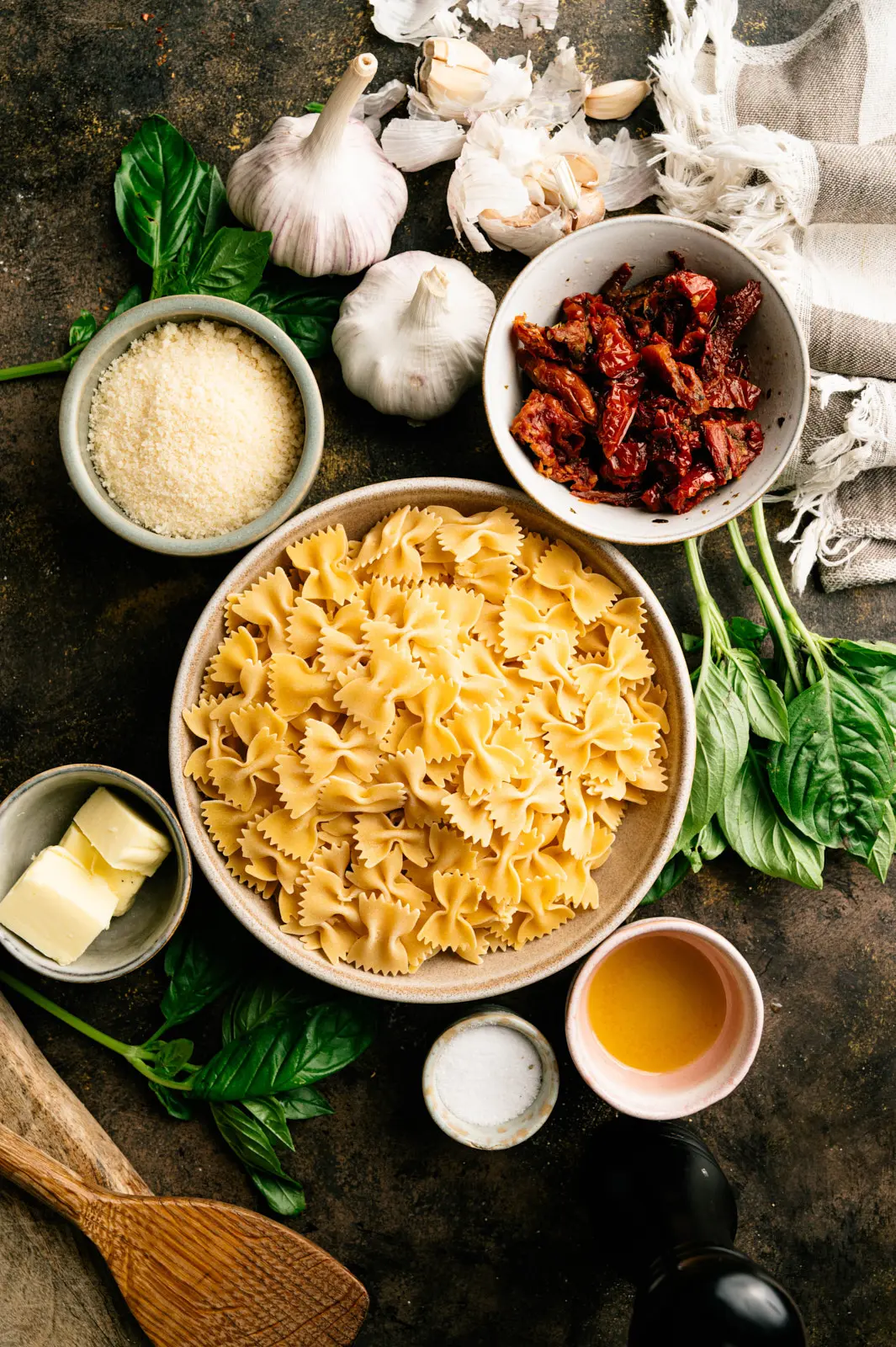 Dry farfalle, sun-dried tomatoes, garlic, basil, butter and grated cheese arranged in bowls on a dark surface.