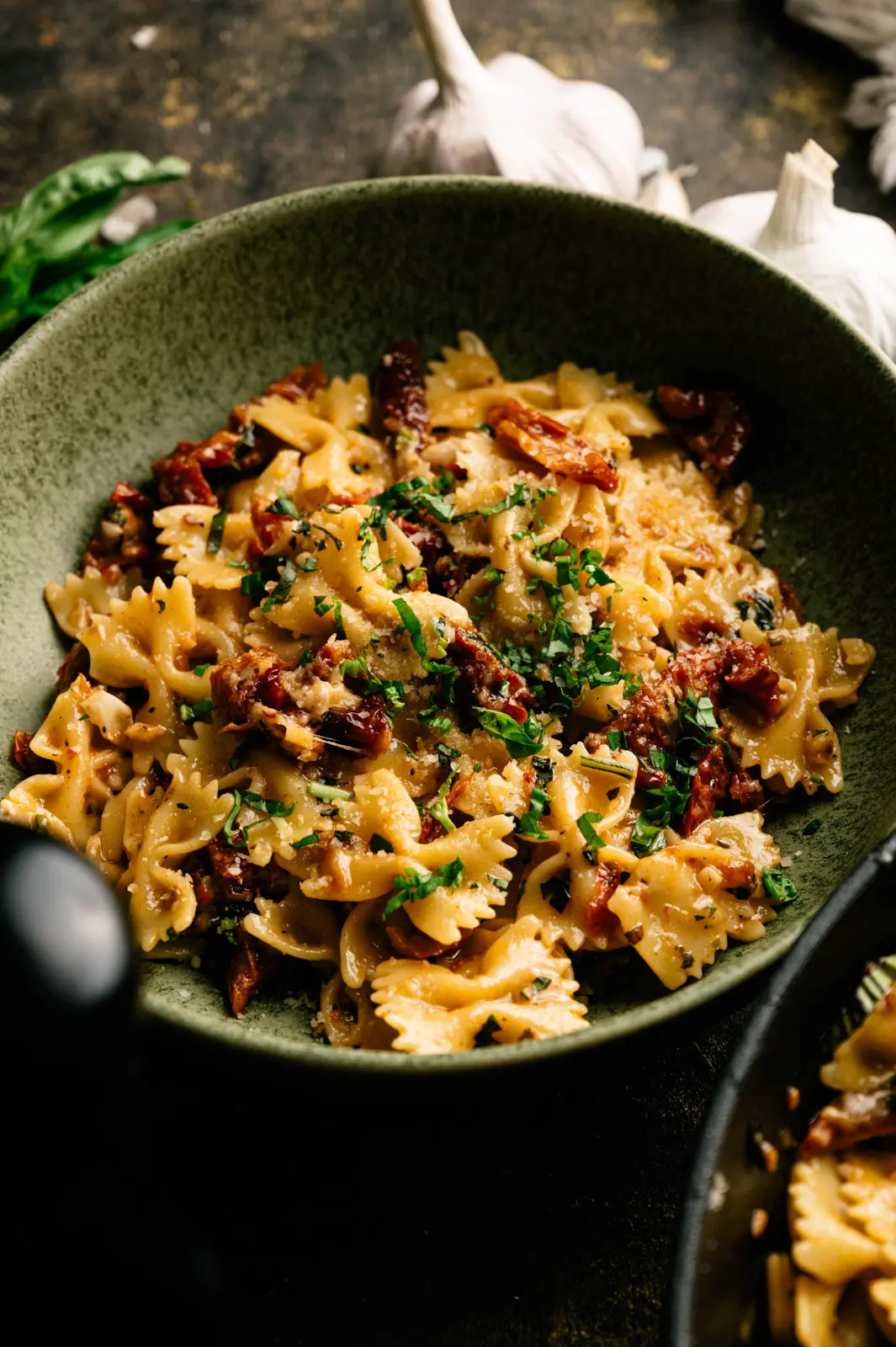 Sun-dried tomato and basil farfalle with grated cheese in a green ceramic bowl, with garlic in the background.