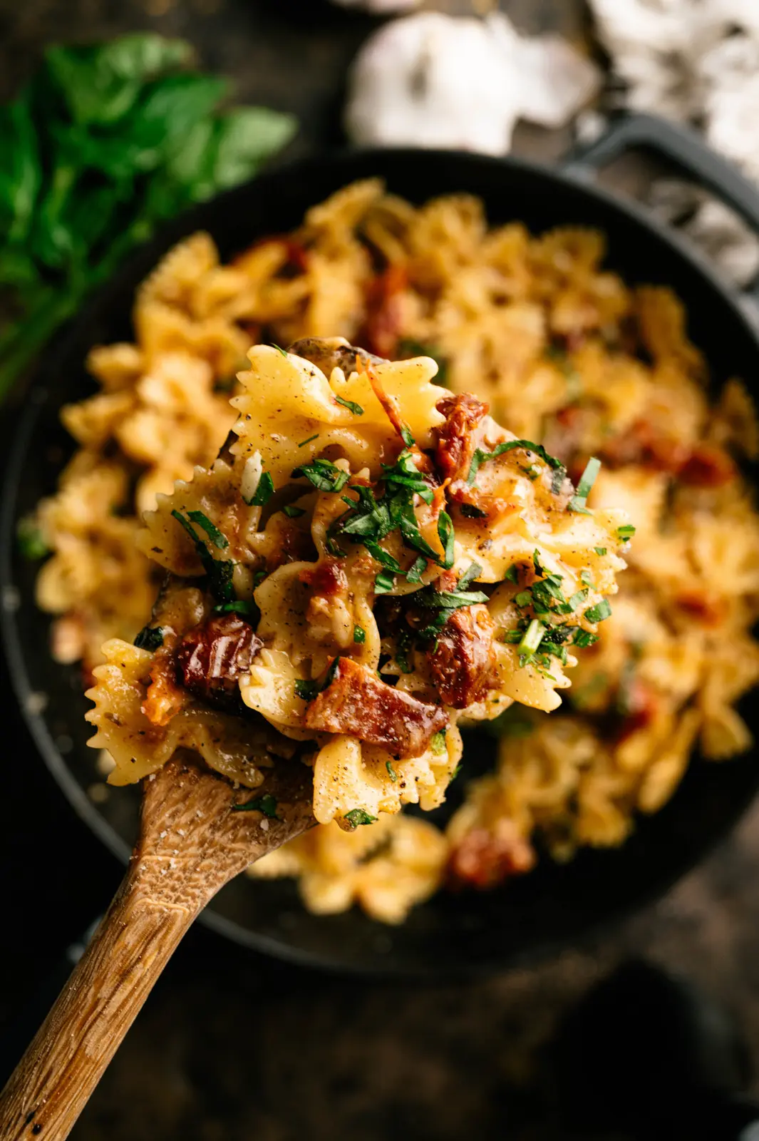 A wooden spoon holds farfalle, sun-dried tomatoes and chopped basil above a softly blurred skillet of pasta.